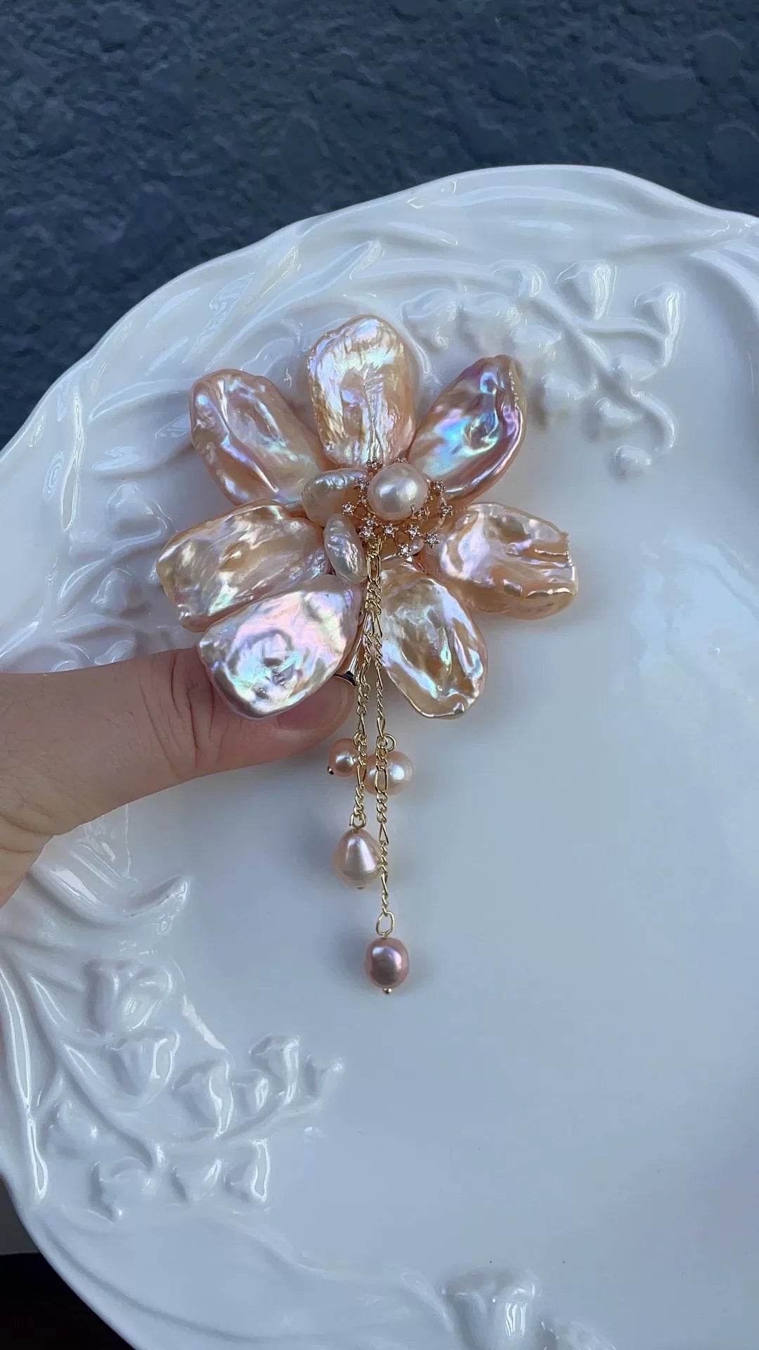 A finger supports a peach-pink flower-shaped ornament with glossy petals and fine gold-colored chains on an embossed plate.