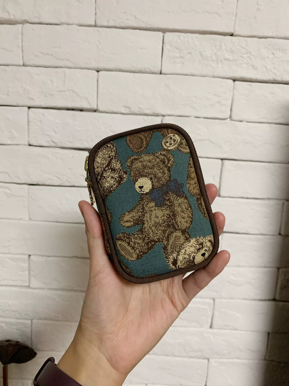 Small rounded rectangular case with a brown teddy bear on dark teal, dark binding around edge.