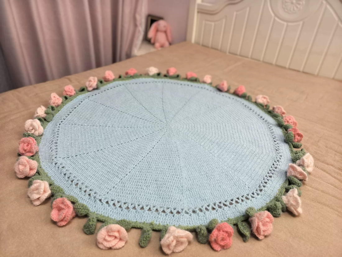 Circular pale blue crochet blanket on a bed with radiating stitch lines and pink and cream flowers around the edge