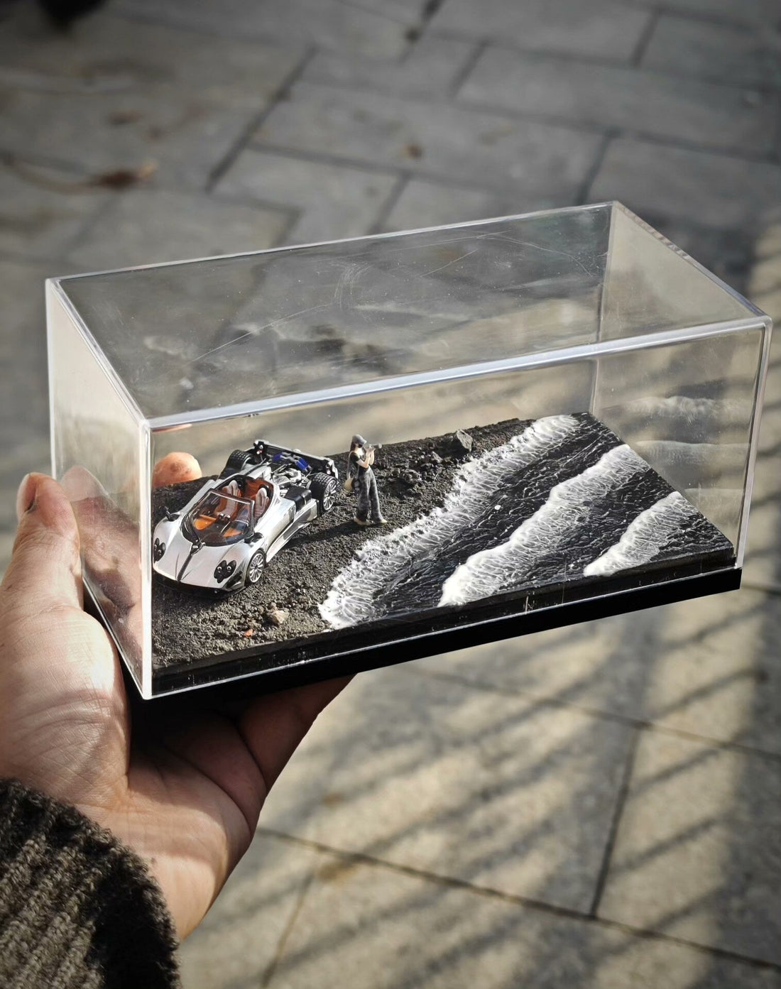 A clear acrylic case holds black sand, parallel white foam waves, a white model car, a dark small vehicle and a tiny standing figure.