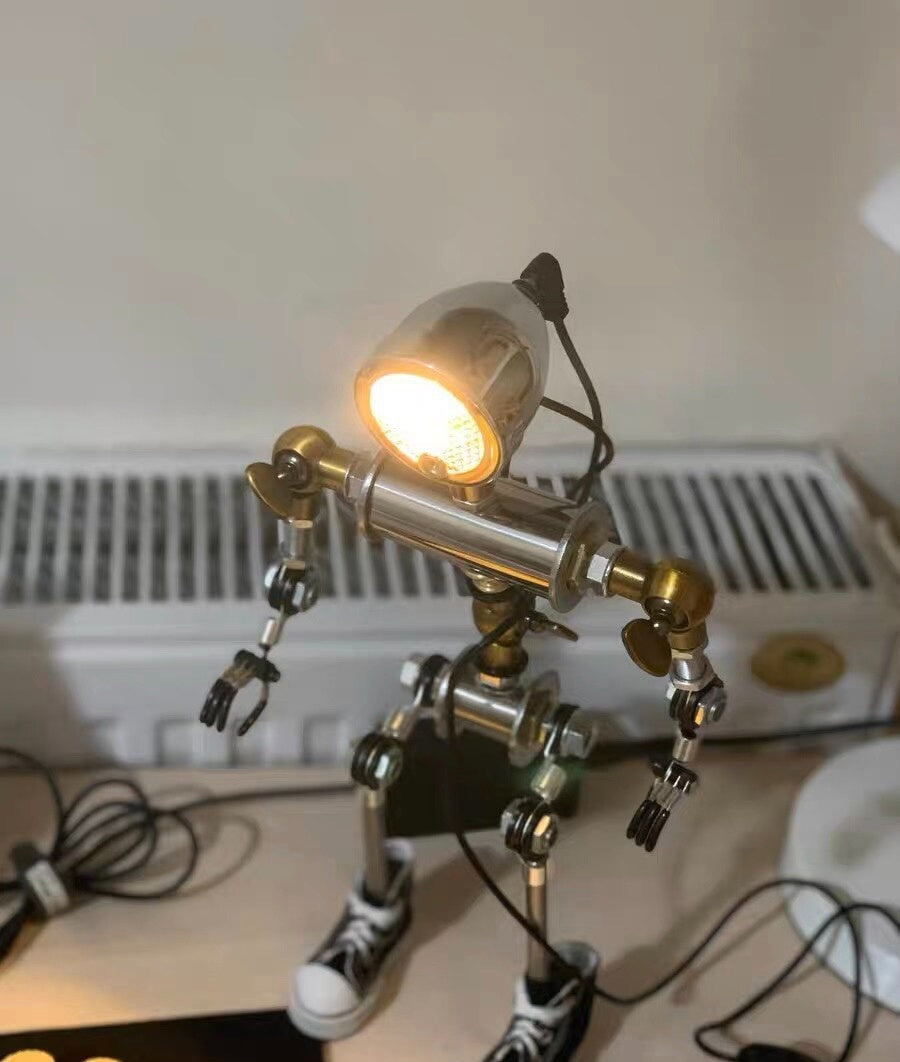 Humanoid metal robot lamp with a glowing rounded head, jointed arms and legs, and shoe-shaped feet